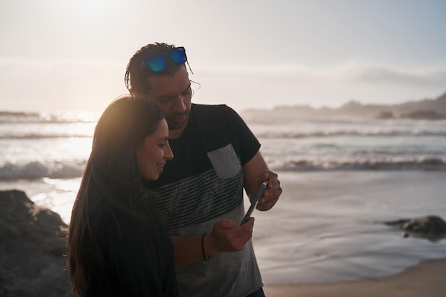 Jeune couple latin sur la plage en regardant le téléphone portable au coucher du soleil