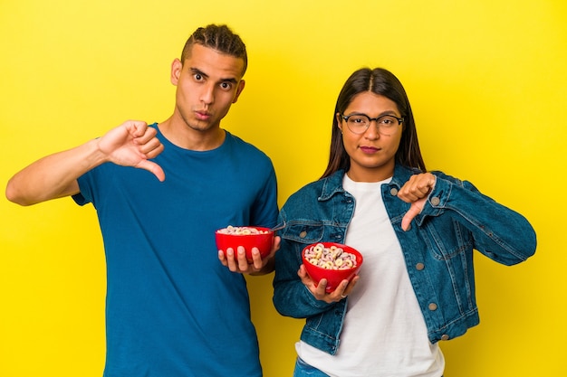 Jeune couple latin tenant un bol de céréales isolé sur fond jaune montrant un geste d'aversion, les pouces vers le bas. Notion de désaccord.