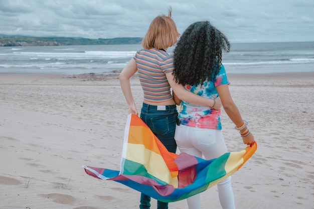 Jeune couple de lesbiennes déplaçant le drapeau de la fierté gay sur une plage de sable tout en regardant un coucher de soleil romantique - Image