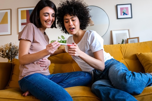 Jeune couple de lesbiennes multiraciales regardant un test de grossesse positif ensemble dans le salon de la maison