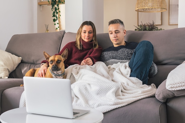 Jeune Couple Et Leur Chien à La Maison Confortable, Se Détendre Sur Le Canapé Tout En Regardant Des Films Sur Ordinateur Portable