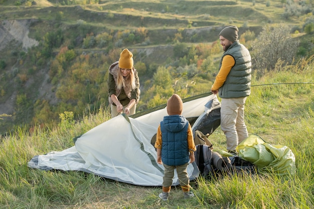 Jeune couple et leur petit fils debout sur l'herbe verte au sommet de la montagne tout en mettant la tente sur le sol devant la caméra