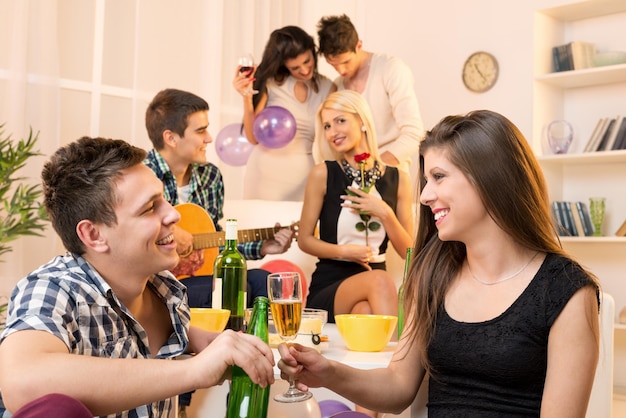 Jeune couple lors d'une fête à la maison, assis par terre et frappant avec des boissons, en arrière-plan, vous pouvez voir leurs amis, deux couples.