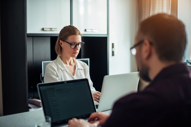 Jeune couple marié adulte assis à une table dans la cuisine, travaillant à distance sur des ordinateurs portables, discutant affaires et souriant. Concentrez-vous sur la belle femme avec des lunettes. L'homme s'assoit à la caméra.