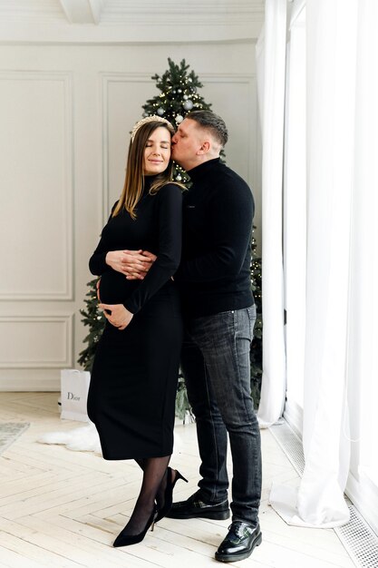 Un jeune couple marié s'embrasse près de l'arbre de Noël dans la maison. une femme enceinte est doucement pressée contre son mari. les futurs parents attendent un bébé. futur enfant. famille enceinte.