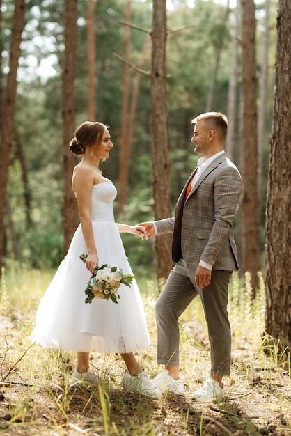 Jeune couple mariée en robe courte blanche et marié en costume gris dans une forêt de pins