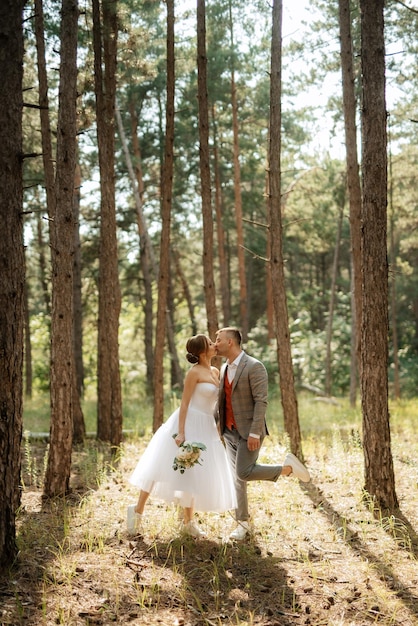 Jeune couple mariée en robe courte blanche et marié en costume gris dans une forêt de pins