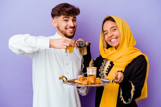 Jeune couple marocain buvant du thé célébrant le mois du ramadan isolé sur mur violet