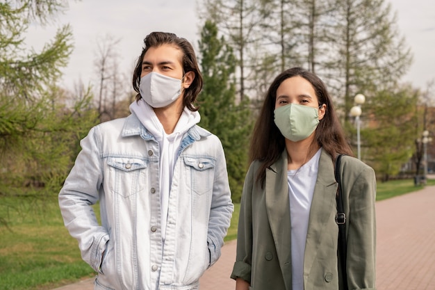 Jeune couple en masques faciaux marchant ensemble dans le parc de la ville pendant l'épidémie de coronavirus