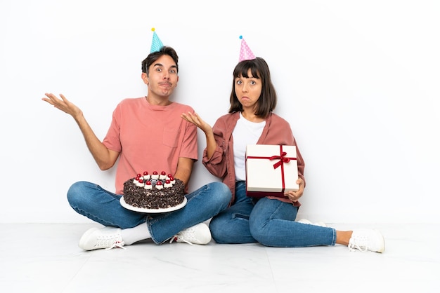 Jeune couple métis célébrant un anniversaire assis sur le sol isolé sur fond blanc ayant des doutes tout en levant les mains et les épaules