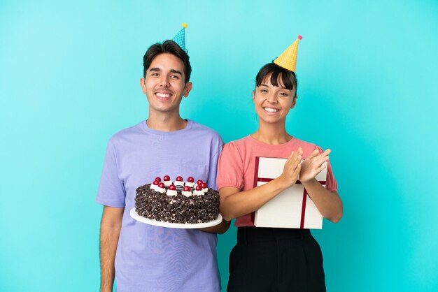 Jeune couple métis tenant un gâteau d'anniversaire et un cadeau isolé sur fond bleu applaudissant après présentation lors d'une conférence