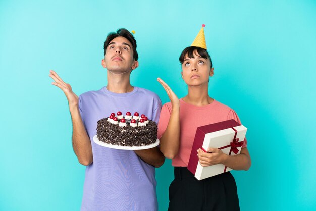 Jeune couple métis tenant un gâteau d'anniversaire et un cadeau isolé sur fond bleu frustré par une mauvaise situation