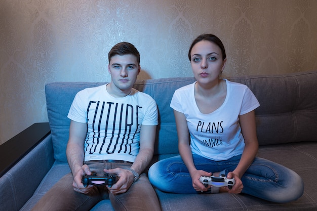 Jeune couple à la mode assis sur un canapé ensemble à la maison jouant à des jeux vidéo avec des expressions compétitives intenses