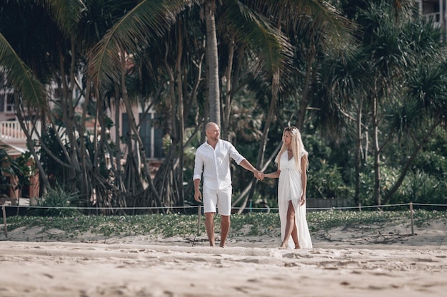 Un jeune couple à la mode séduisant en vêtements blancs se promène sur la plage et se tient la main. Belle blonde aux cheveux longs et un homme chauve sont en vacances. Phuket. Thaïlande.