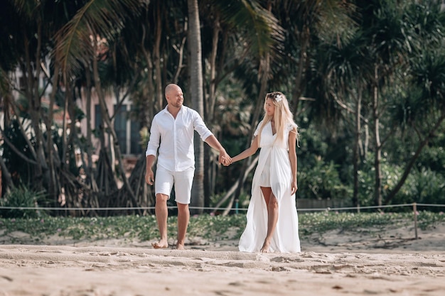 Un jeune couple à la mode séduisant en vêtements blancs se promène sur la plage et se tient la main. Belle blonde aux cheveux longs et un homme chauve sont en vacances. Phuket. Thaïlande.