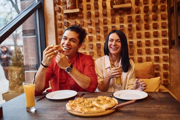 Jeune couple multiethnique assis ensemble à l'intérieur et mangeant de la pizza.