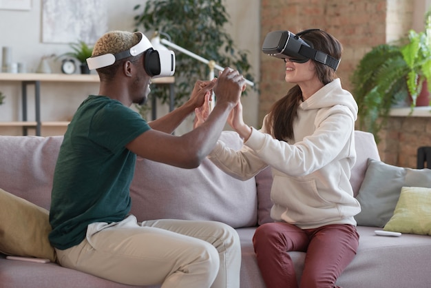 Jeune couple multiethnique dans des lunettes de réalité virtuelle se tenant la main en jouant à un jeu vidéo assis sur un canapé dans la chambre