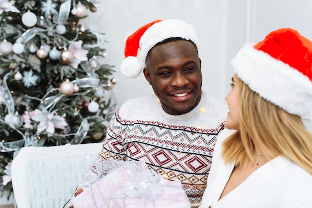 Un jeune couple multiethnique échange des cadeaux dans un emballage rose. Un homme noir et une femme de race blanche sont assis sur un canapé dans un intérieur de Noël clair