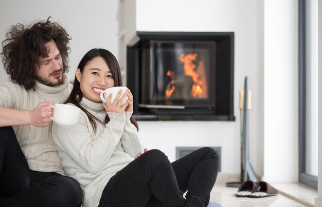 Jeune couple multiethnique romantique assis sur le sol devant la cheminée à la maison, parlant et buvant du café lors d'une froide journée d'hiver