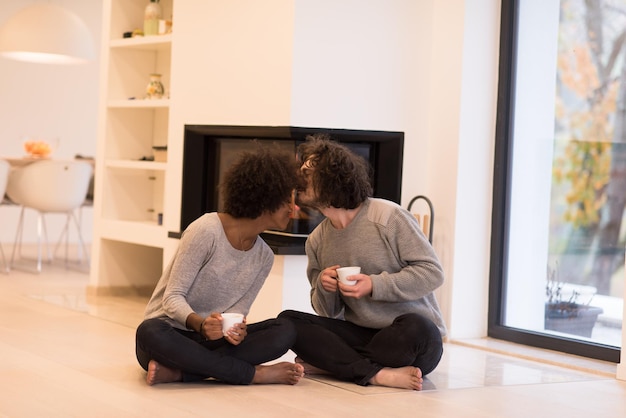 Jeune couple multiethnique romantique assis sur le sol devant la cheminée à la maison, se regardant, parlant et buvant du café le jour de l'automne