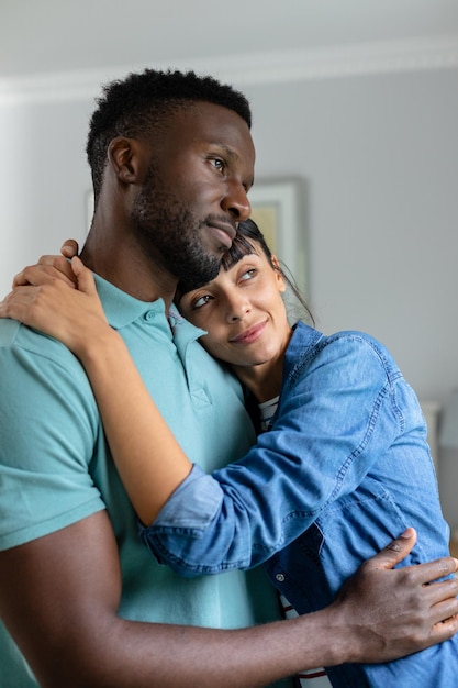 Photo un jeune couple multiracial réfléchi regarde ailleurs tout en s'embrassant dans le salon à la maison. inchangé, mode de vie, vie domestique, convivialité, amour, contemplation.