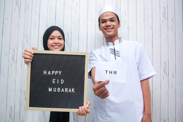 Photo jeune couple musulman tenant une enveloppe blanche appelée thr tunjangan hari raya et un tableau aux lettres dit joyeux eid mubarak