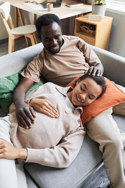 Un jeune couple noir attend un bébé et se câline sur le canapé.