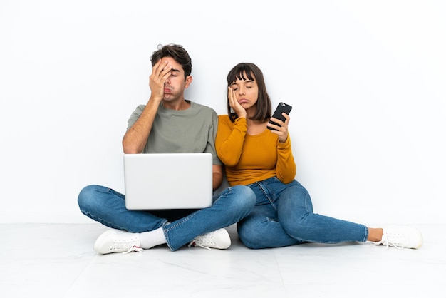 Jeune couple avec un ordinateur portable et un mobile assis sur le sol avec une expression faciale surprise et choquée