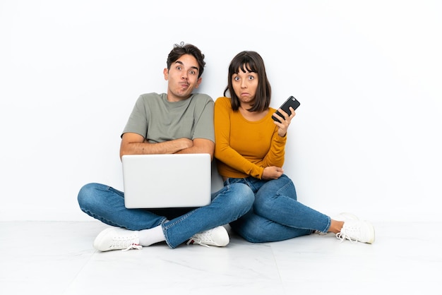 Jeune couple avec un ordinateur portable et un mobile assis sur le sol faisant un geste de doute tout en levant les épaules