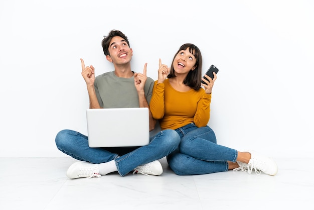 Jeune couple avec un ordinateur portable et un mobile assis sur le sol pointant avec l'index une excellente idée
