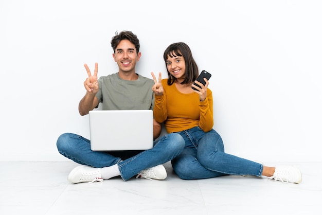 Jeune couple avec un ordinateur portable et un téléphone portable assis sur le sol souriant et montrant le signe de la victoire