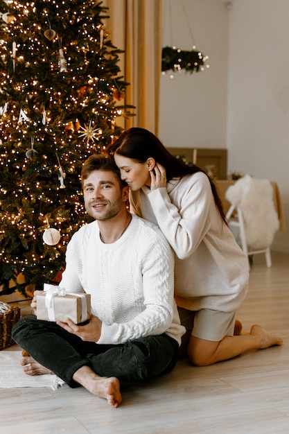 Jeune couple ouvrant des cadeaux de Noël dans leur salon en face de l'arbre de Noël