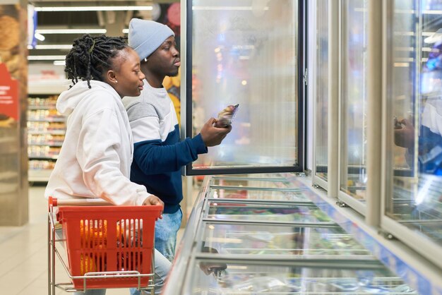 Jeune couple avec panier choisissant des produits alimentaires surgelés au supermarché