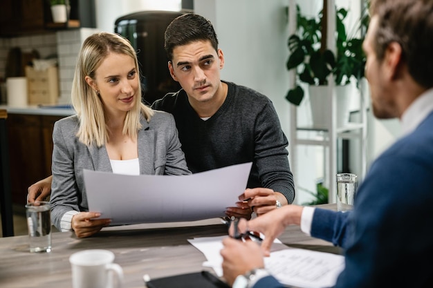 Jeune couple parlant avec un agent immobilier tout en analysant des plans de logement pendant la réunion