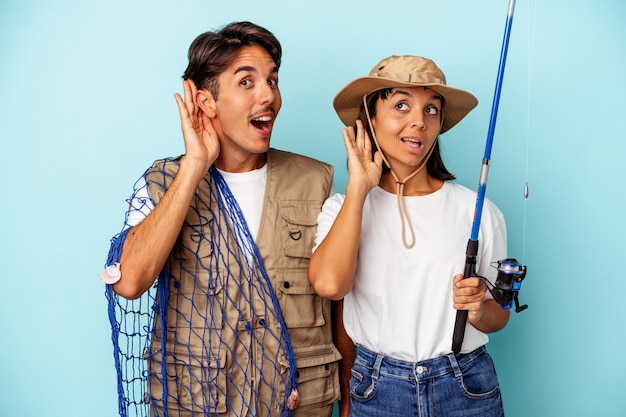 Jeune couple de pêcheurs de race mixte isolé sur fond bleu essayant d'écouter un potin.