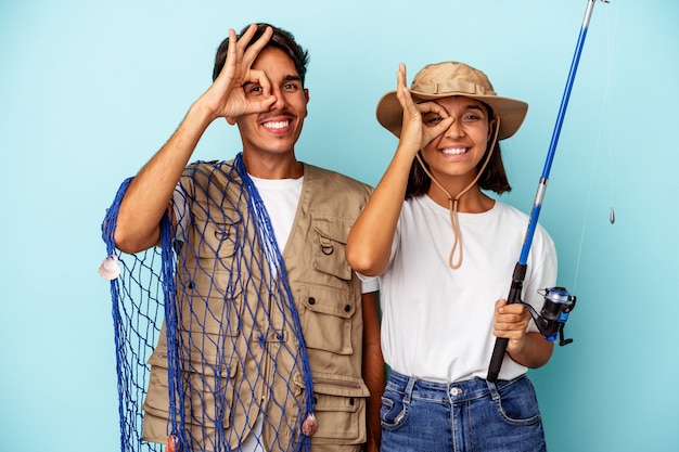 Jeune couple de pêcheurs de race mixte isolé sur fond bleu excité en gardant le geste ok sur les yeux.