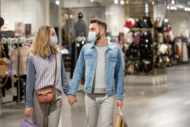 Jeune couple portant des masques de protection se tenant la main en marchant dans le magasin de vêtements