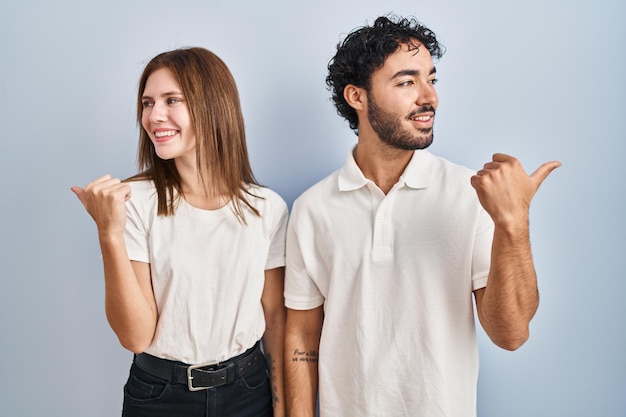 Jeune couple portant des vêtements décontractés debout ensemble souriant avec un visage heureux regardant et pointant vers le côté avec le pouce vers le haut.