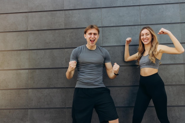 Jeune couple portant des vêtements de sport debout sur le mur à l'extérieur célébrant la folie et étonné du succès avec les bras levés et les yeux ouverts criant le concept de gagnant excité