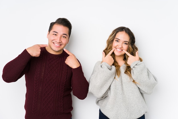 Jeune couple posant dans un fond blanc sourit, pointant les doigts sur la bouche.