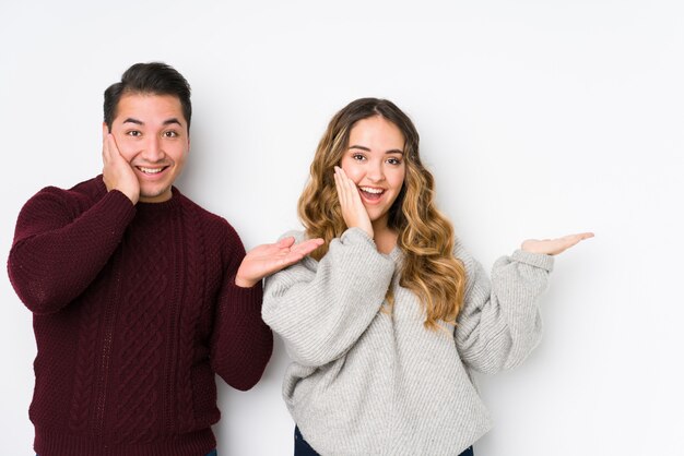 Jeune couple posant dans un mur blanc détient un espace de copie sur une paume, gardez la main sur la joue. Etonné et ravi.