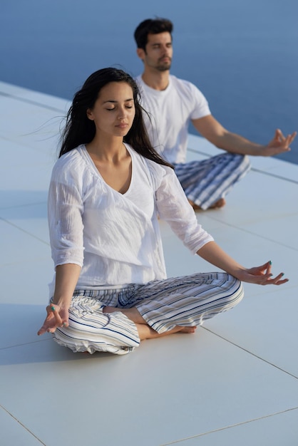 jeune couple pratiquant le yoga au coucher du soleil dans une terrasse de maison moderne avec l'océan et le coucher du soleil en arrière-plan