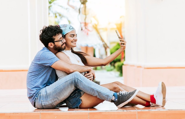 Jeune couple prenant un selfie assis sur le sol Jeune couple latin amoureux assis sur le sol prenant un selfie à l'extérieur Concept de couple heureux prenant des photos avec le téléphone portable