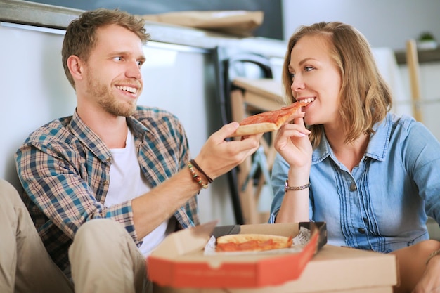 Un jeune couple prend une pause déjeuner pizza sur le sol après avoir emménagé dans une nouvelle maison avec des boîtes autour d'eux