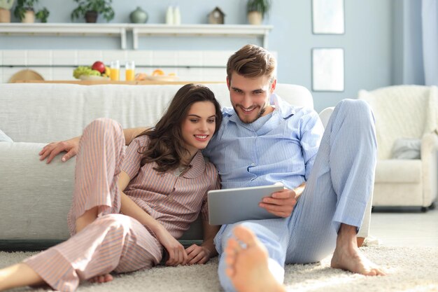 Jeune couple en pyjama regardant du contenu multimédia en ligne sur une tablette assise par terre dans le salon.