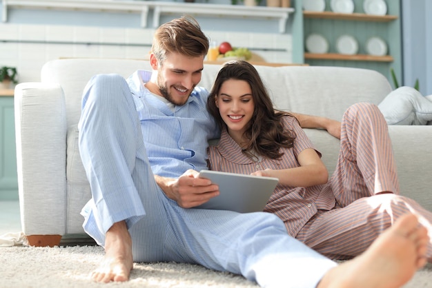 Jeune couple en pyjama regardant du contenu multimédia en ligne sur une tablette assise par terre dans le salon.