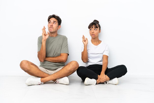 Jeune couple de race mixte assis sur le sol isolé sur fond blanc avec les doigts croisés et souhaitant le meilleur