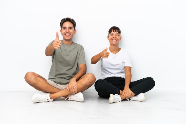 Jeune couple de race mixte assis sur le sol isolé sur fond blanc donnant un geste du pouce parce que quelque chose de bien s'est produit