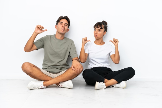 Jeune couple de race mixte assis sur le sol isolé sur fond blanc fier et satisfait de l'amour vous-même concept