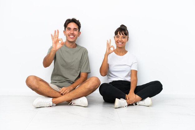 Jeune couple de race mixte assis sur le sol isolé sur fond blanc montrant un signe ok avec les doigts